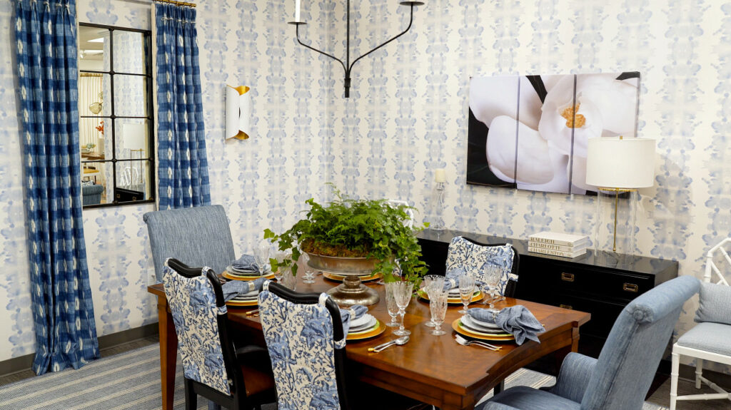 A formal dining room scene with light blue wallpaper, blue curtains, and a wooden dining table with blue upholstered chairs. On the table is a large green plant, crystal wine glasses and white and gold plate settings. 