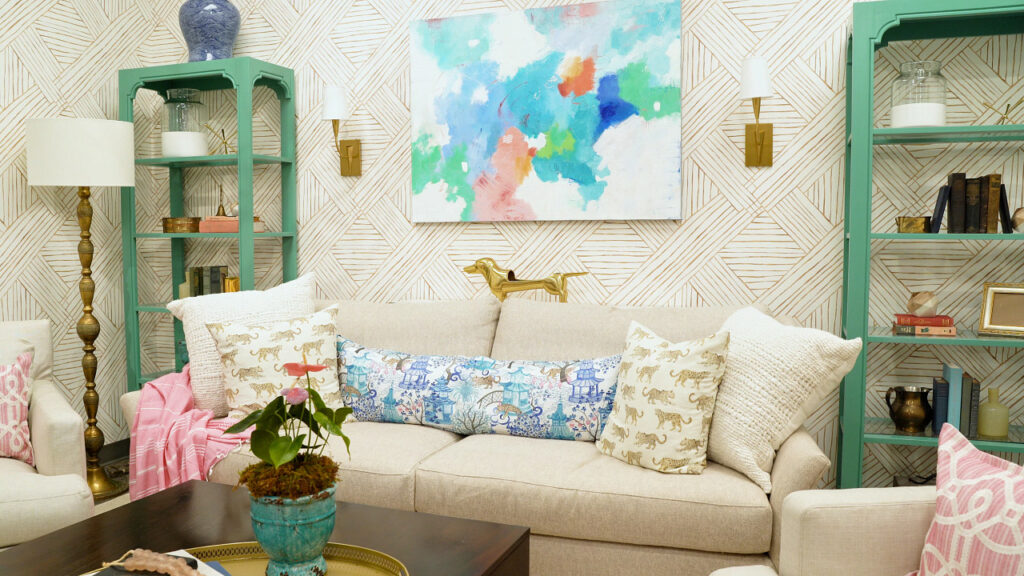 Living room scene with gold, geometric woven wallpaper, a beige couch with a gold weiner dog figurine behind it, and blue and beige chinoiserie-inspired pillows with leopards on them. There is a gold lamp and green bookshelves against the wall.