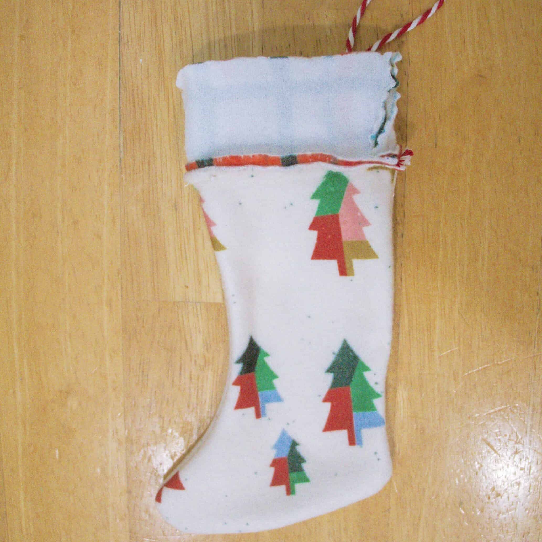An almost completed mini stocking lays on a wooden surface. The stocking’s boot features a pattern with dark green, Kelly green, light blue and brown color-blocked trees on a cream background. The cuff has been sewn to the boot but has yet to be turned inside the boot. The white “wrong” side of the fabric is still sticking out of the boot’s top. The top of a loop of red-and-white ribbon is sticking out of the stocking’s top right.