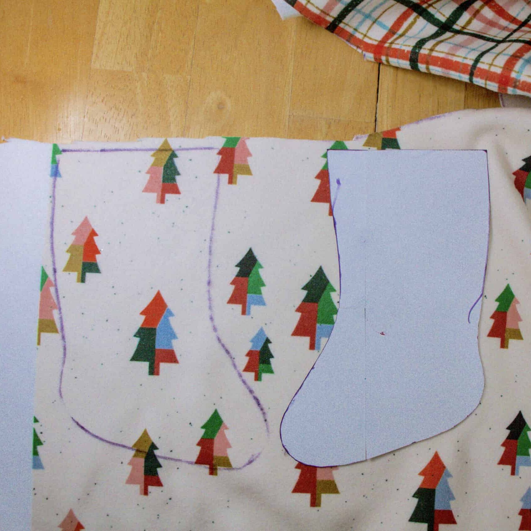 One side of a stocking has been tried onto fabric featuring a design on the fabric with dark green, Kelly green, light blue and brown color-blocked trees on a cream background. The other side of the stocking has not been cut out yet, but the paper pattern piece is laying on top of the fabric.