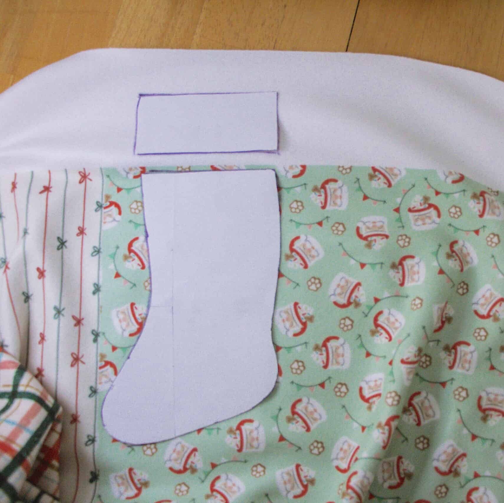 The stocking pattern’s boot and cuff have been cut in white paper. The piece of paper for the stocking’s cuff is laying on top of the white selvage edge of the fabric. The piece of paper for the stocking’s boot is laying on top of mint green fabric with a design of white mugs with red rims and handles with whipped cream and sprinkles on their tops. Small strips of fabric bunting are floating around the design as are white paw prints.