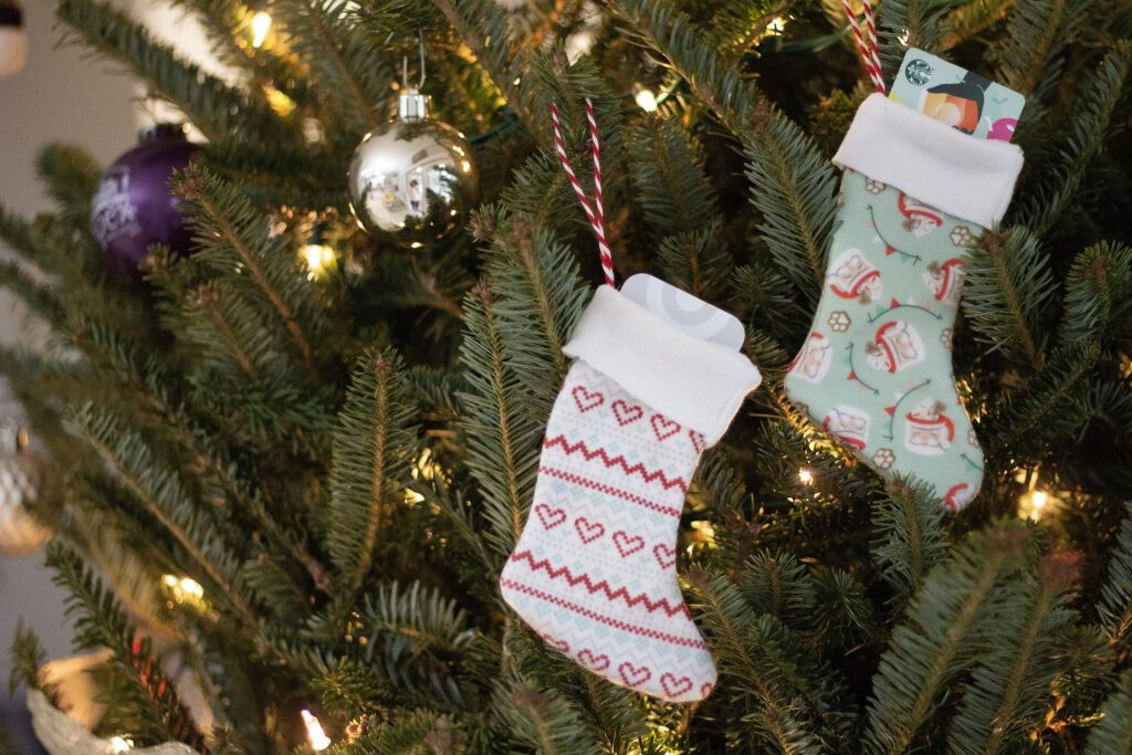 Two mini stocking gift card ornaments hang from a Christmas tree from red-and-white ribbon loops. The stocking to the right has a white cuff and a boot with repeating small white mugs with Santa’s face on them. They have red rims and handles and a snowflake and cookie sticking out of the top. Red, white and green strips of bunting float through the design on a mint background. The stocking to the left has a white cuff and a boot with a blue and red Fair Isle design featuring hearts and zig zag lines. Two round ornaments, one silver, one purple, are to the left of the stockings.