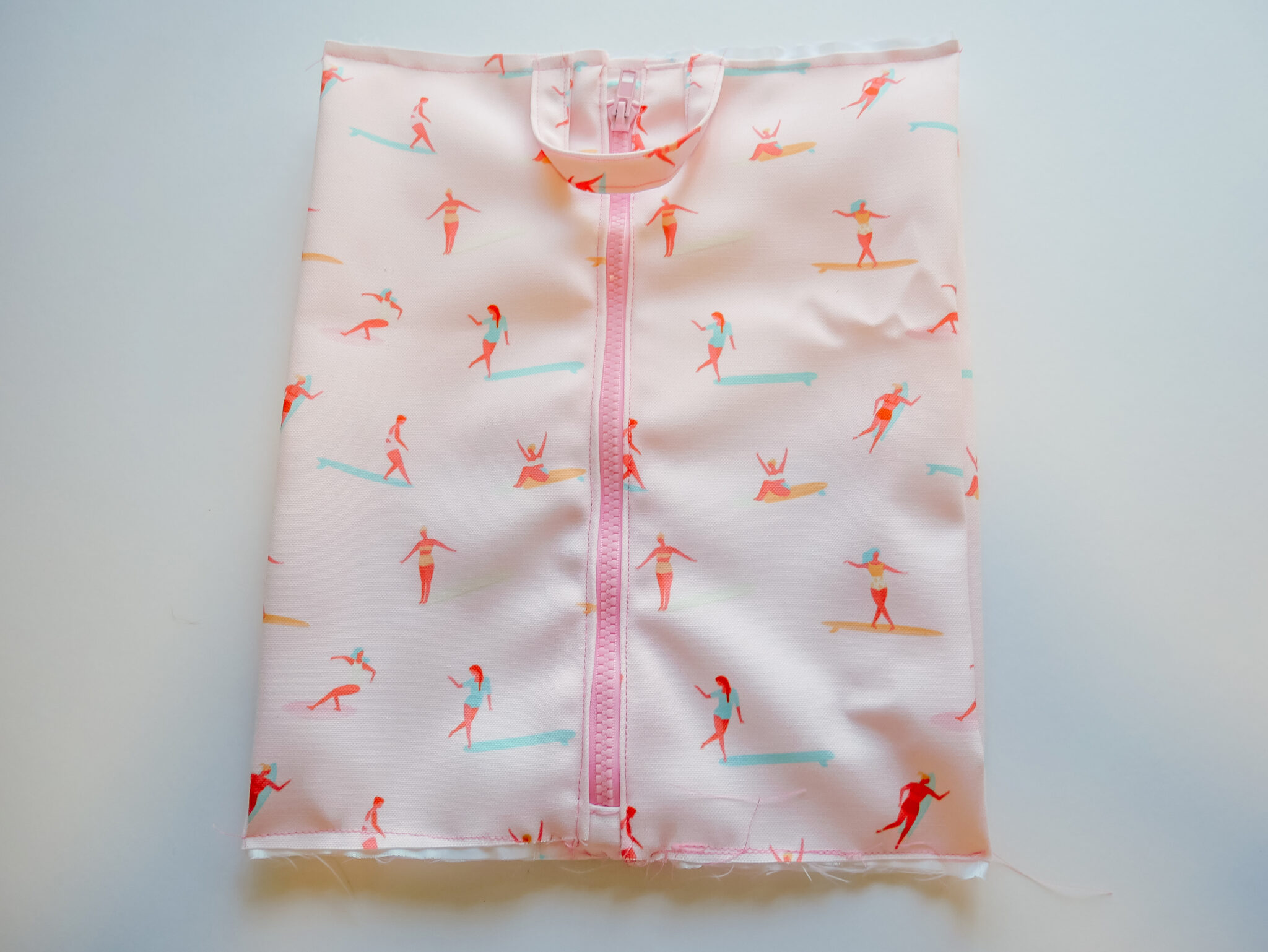 A fabric rectangle featuring a design with small female surfers surfing through a pink background lays on a white surface. A pink zipper has been stitched up the middle. A U-shaped handle in the same fabric has been sewn to the top center of the bag. The bag is laying on a white surface.