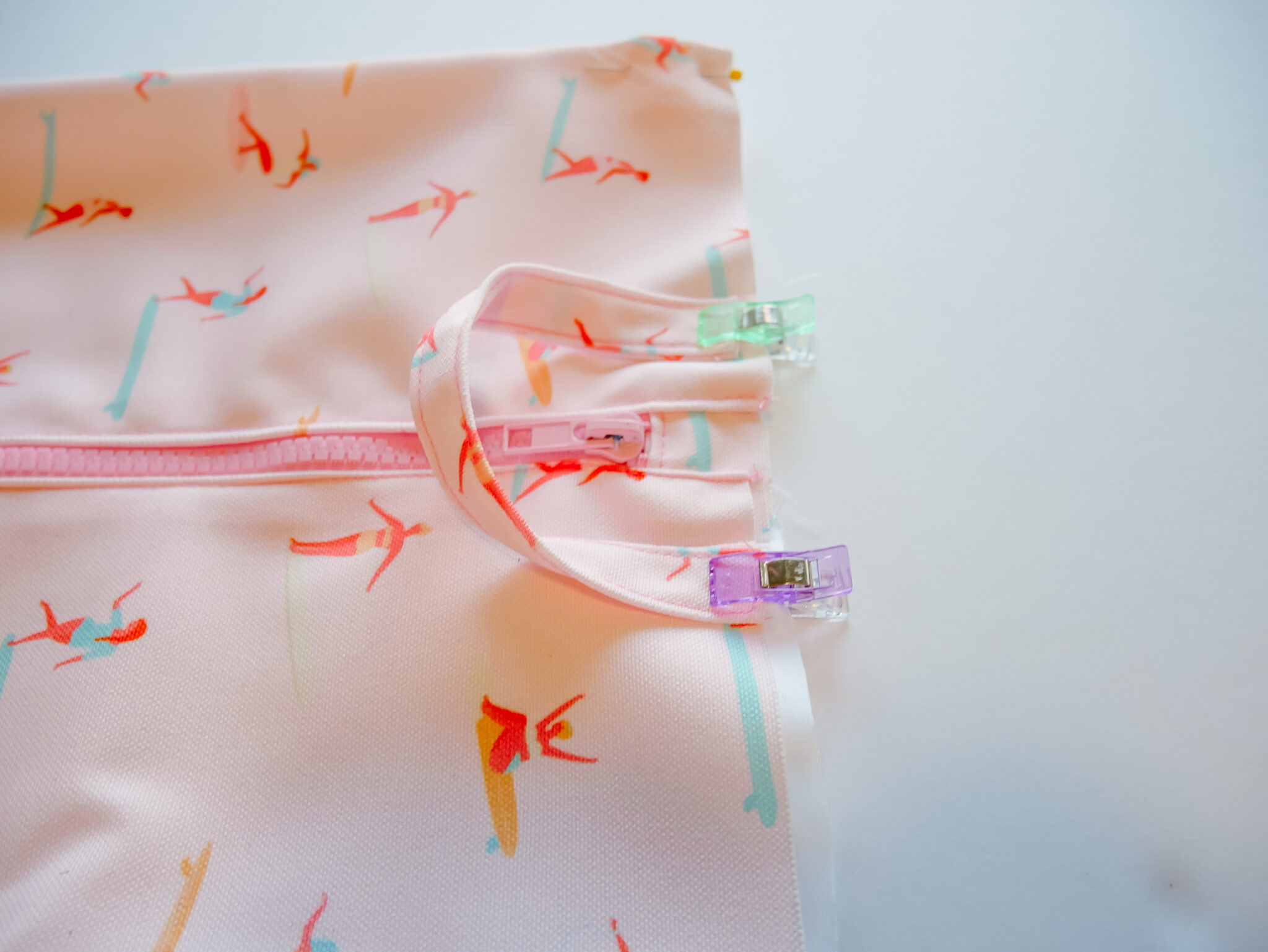 A fabric rectangle featuring a design with small female surfers surfing through a pink background lays on a white surface. A pink zipper has been stitched up the middle. A U-shaped handle in the same fabric has been placed to the center right of the rectangle and is being held together by small clips.