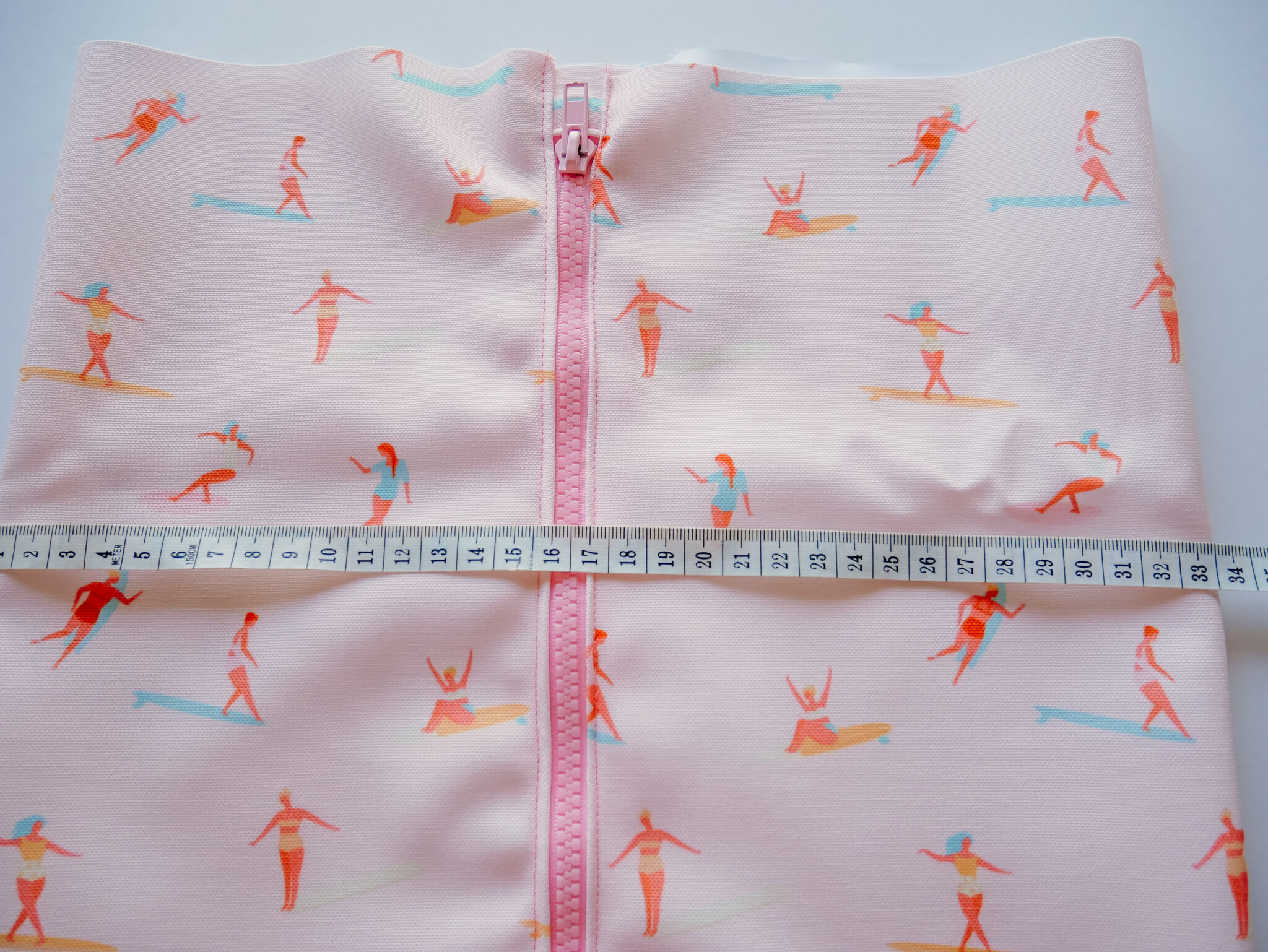 A fabric rectangle featuring a design with small female surfers surfing through a pink background lays on a white surface. A pink zipper has been stitched up the middle. A small white measuring tape has been placed over the fabric.