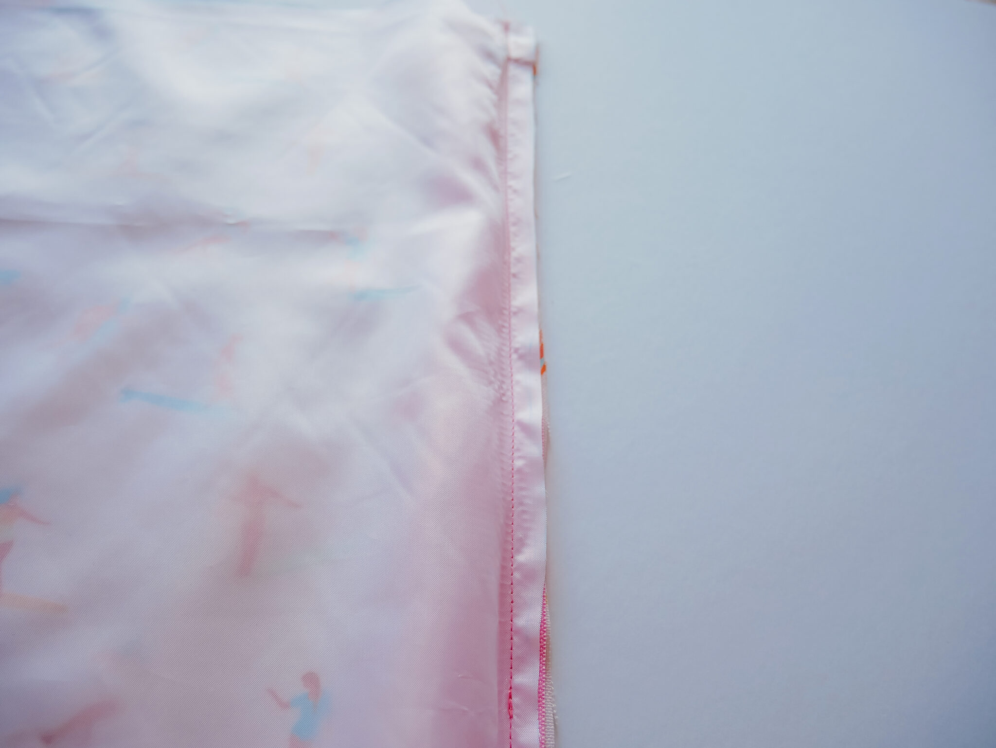 A rectangle piece of Polyurethane Laminate fabric is lying on top of a rectangle of fabric featuring a design with small female surfers surfing through a pink background. A pink zipper lays in between the two. The right edge of those three things has been sewn together.