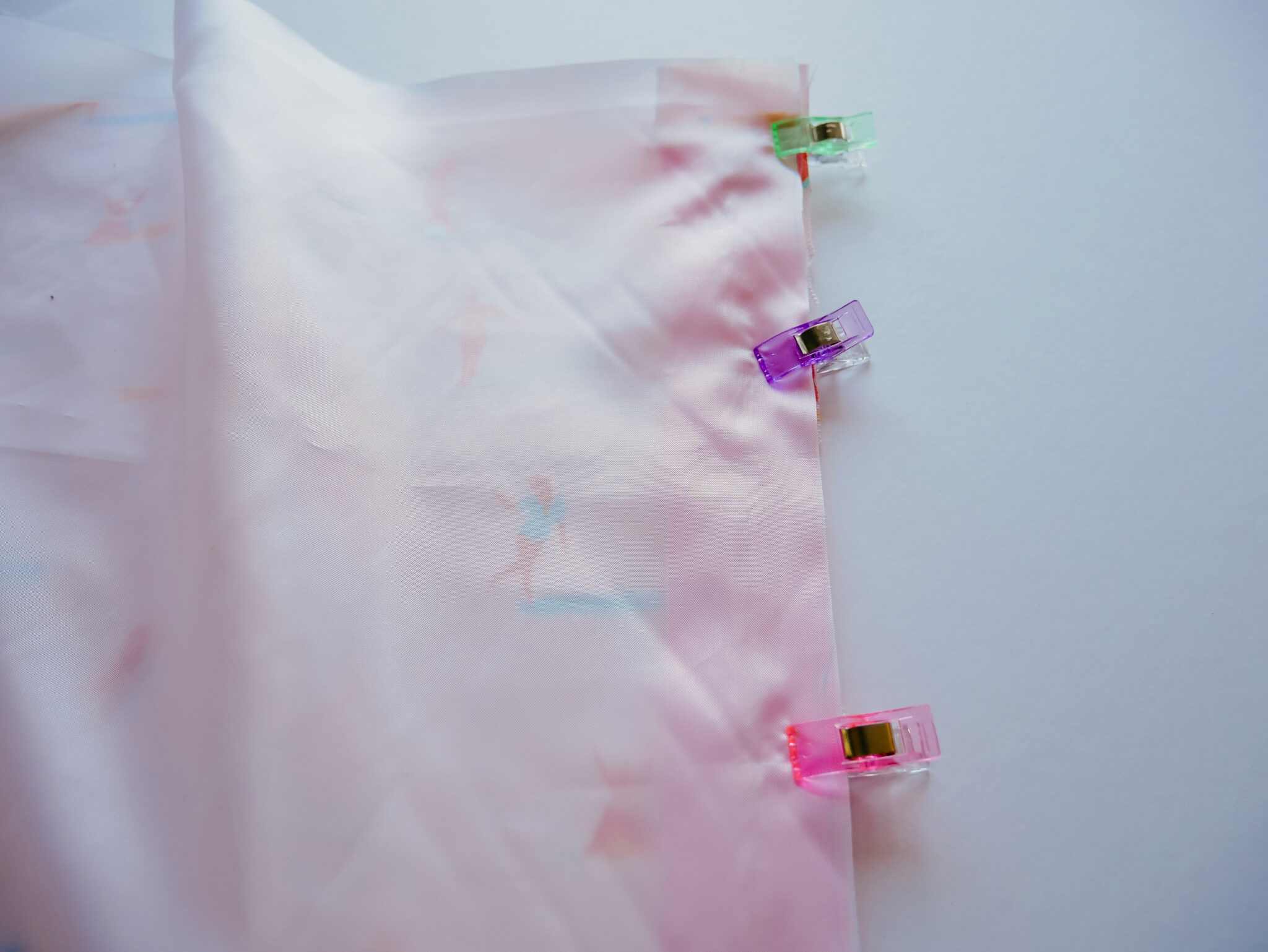 A rectangle piece of Polyurethane Laminate fabric is lying on top of a rectangle of fabric featuring a design with small female surfers surfing through a pink background. A pink zipper lays in between the two. Pastel wonder clips have been placed along the right edge in order to sew those three things together.