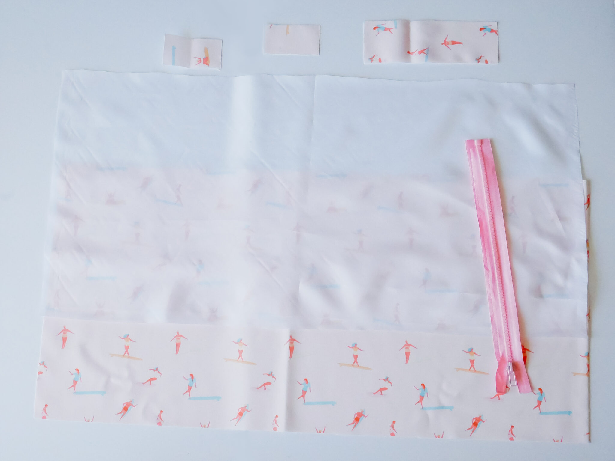 Two rectangle pieces of fabric for the wet bag are lying on a white surface. The piece on top has been cut out of Polyurethane Laminate. The piece on the bottom has a design featuring small female surfers surfing through a pink background. Three smaller rectangles in the pink fabric are at the top of image, two small rectangles to the left and one larger rectangle to the right. A pink zipper is lying on top of the fabric to the right.