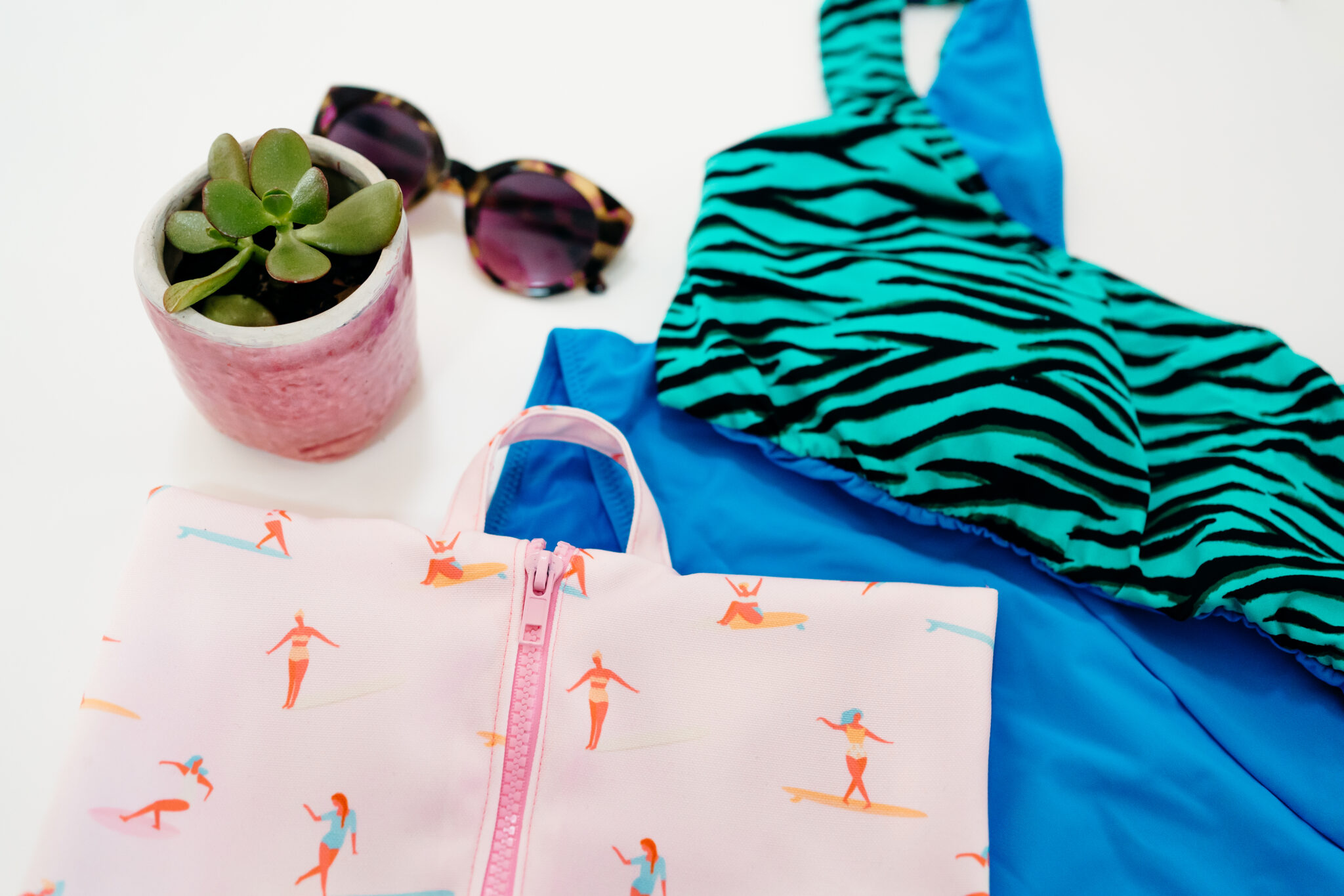 A fabric rectangle featuring a design with small female surfers surfing through a pink background lays on a white surface. A pink zipper has been stitched up the middle. A U-shaped handle in the same fabric has been sewn to the top center of the bag. The bag is laying on a white surface and to the left of a teal-and-black swimsuit and blue wetsuit. A small succulent is to the top left along with tortoiseshell sunglasses.