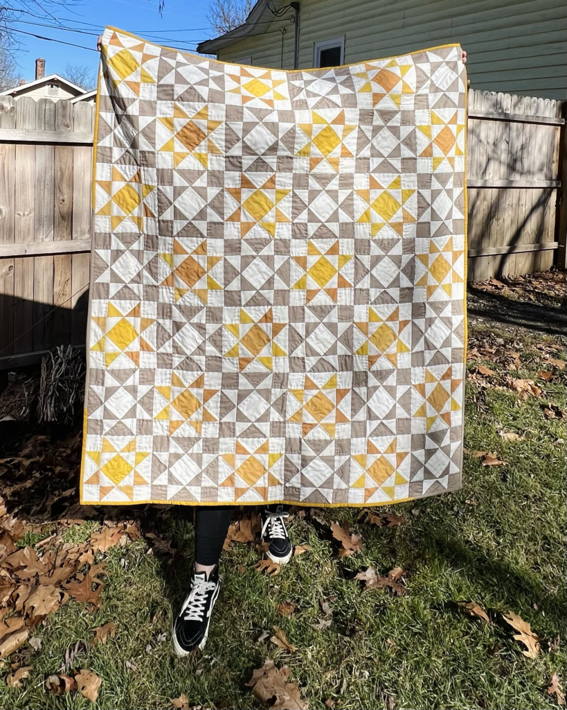 Kadie holds up a white, brown and yellow quilt with a star motif. In some places, the yellow bleeds a bit into orange. Kadie’s feet are visible at the bottom of the photo and they are wearing black-and-white shoes. They are standing in a patch of green grass with some large brown leaves and in front of a brown wooden gate.