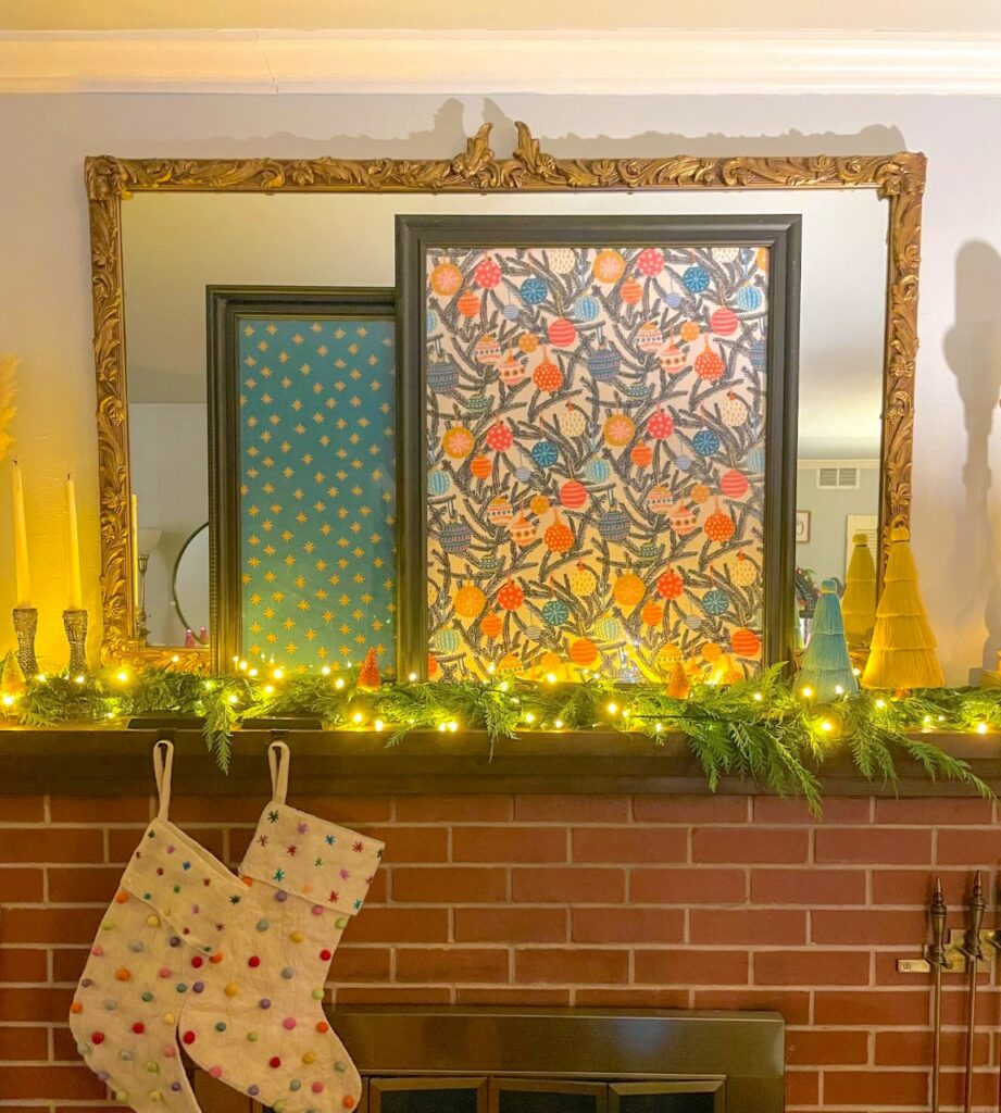 A red brick fireplace has two gray stockings covered in colorful baubles hanging from a dark brown mantel on its lefthand side. Above the fireplace is a large rectangular mirror. Leaning against the mirror are two pieces of Spoonflower fabric in black frames. The back frame shares a design with a teal background and gold stars. The front frame is of a Christmas tree in dark blue, with colorful ornaments hanging all over its branches.