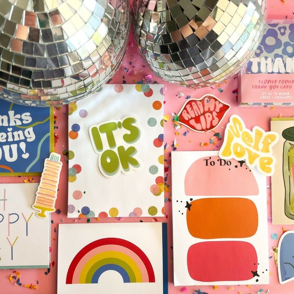 Two disco balls sit at the top of an image with several different designs on notecards stickers and paper below. A sticker that says “It’s ok” sits on top of a notepad with colorful round jewel tone dots around the edge. Stickers that say “read my lips” and “self love” are peppered in with a to-do notepad with geometric ombre blobs, a rainbow notecard.