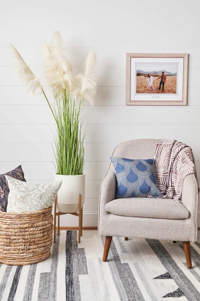 A gray chair sits to the right, with a light blue pillow with a white geometric design on it and a burgundy-and-white blanket draped over it. A tall planter of sea oats is to the middle of the photo with a basket of neutral pillows to the left.