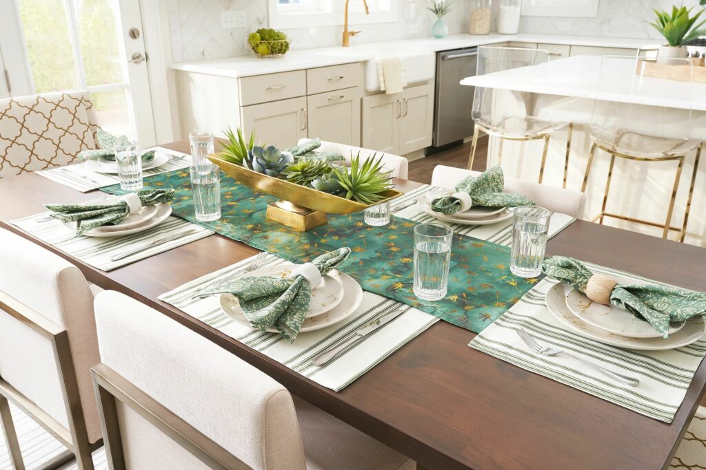 A wooden kitchen table has white and green striped placemats at each of the six table settings. A jade green table runner runs down the middle of the table. On top of the white ceramic plates are green napkins folded in wooden napkin rings. At the center of the table is a long gold planter with succulents.