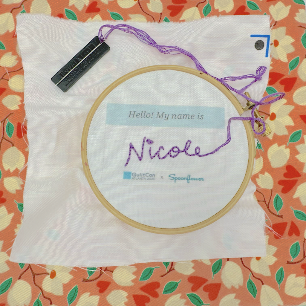 An embroidered name tag that has “Hello! My name is:” printed at the top and says “QuiltCon” and “Spoonflower” printed at the bottom sits finished in a wooden embroidery hoop. The name Nicole is in cursive and stitched in purple embroidery floss. The “i” in Nicole is stitched in purple with a French knot.