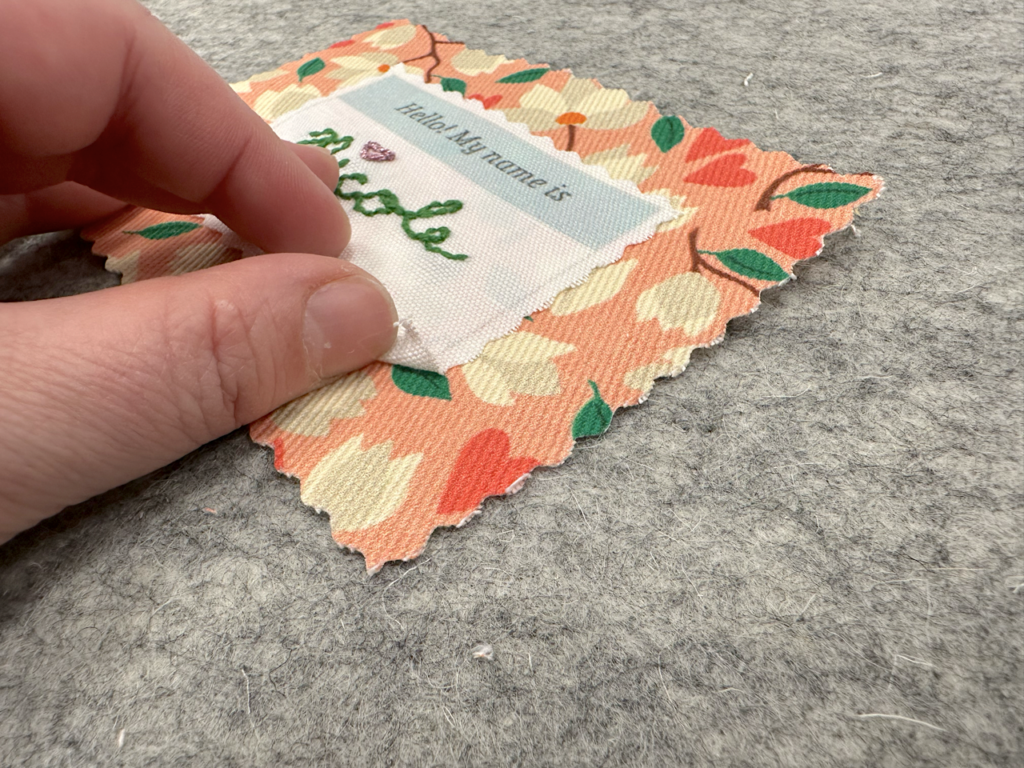 A name tag has been embroidered in green floss with a red heart over the “i.” It says “Hello! My name is:” at the top and says “QuiltCon” and “Spoonflower” at the bottom. The top fabric is white. A larger square of fabric underneath the name tag has a peach background and cream magnolias, pink hearts and green leaves. A hand is checking to see if the name tag has been adhered to the backing fabric with seam tape.