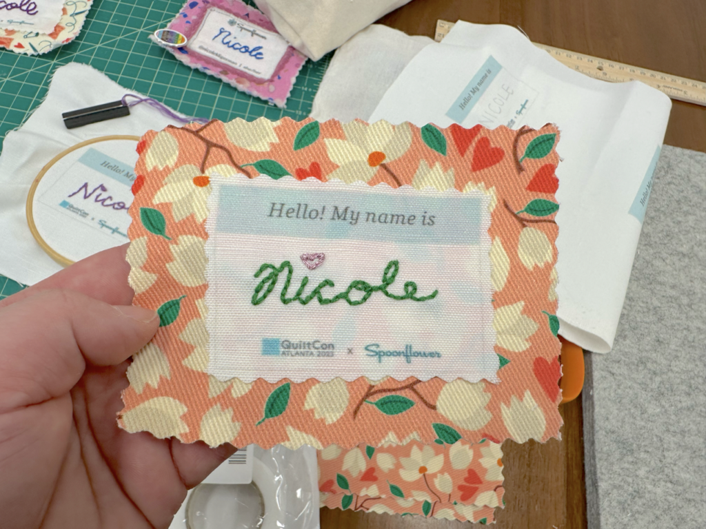 A name tag has been embroidered in green floss with a red heart over the “i.” It says “Hello! My name is:” at the top and says “QuiltCon” and “Spoonflower” at the bottom. The top fabric is white. A larger square of fabric underneath the name tag has a peach background and cream magnolias, pink hearts and green leaves. A hand holds up the finished name tag.