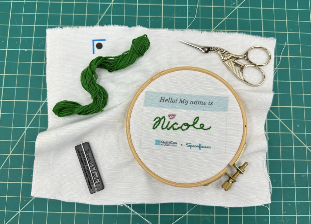 An embroidered name tag that has “Hello! My name is:” printed at the top and says “QuiltCon” and “Spoonflower” printed at the bottom sits finished in a wooden embroidery hoop. The name Nicole in cursive and stitched in green embroidery floss. The “i” in Nicole is stitched in red with a heart.