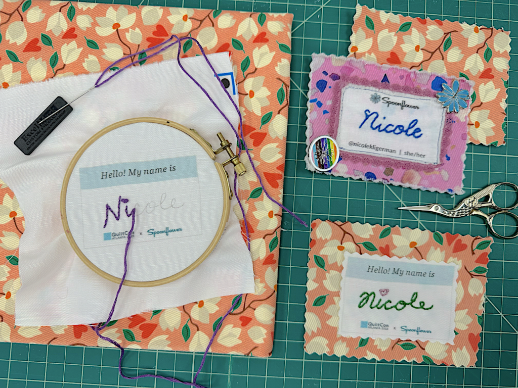 An embroidered name tag that has “Hello! My name is:” printed at the top and says “QuiltCon” and “Spoonflower” printed at the bottom sits finished in a wooden embroidery hoop. The name Nicole is written in cursive in pencil on the canvas. The letters “n” and “i” have been stitched with 6 strands of purple embroidery floss. The “i” in Nicole is stitched in purple with a French knot. Two examples of finished embroidered name tags, one with the name Nicole in cursive and stitched in green embroidery floss. The “i” in Nicole is stitched in red with a heart and one with the name Nicole in cursive and stitched in blue embroidery floss sit to the right.