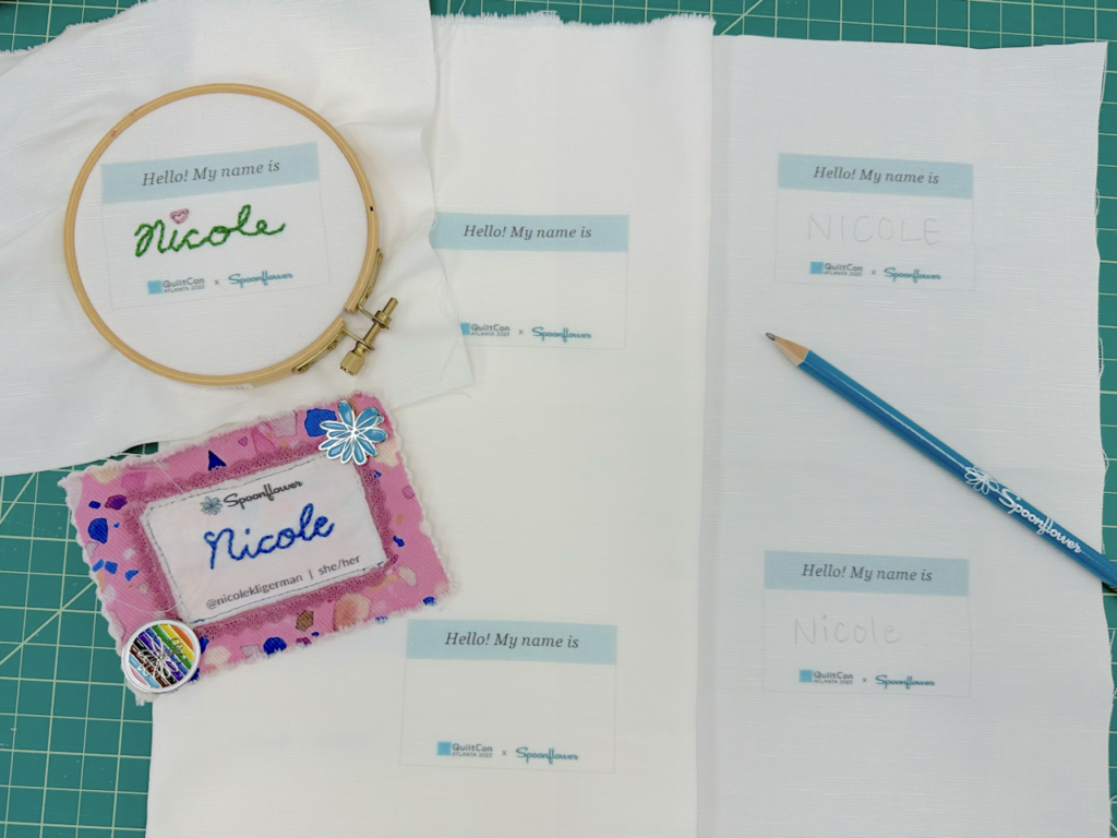 Two embroidered name tags sit to the left of the image, one is purply pink and the other has been embroidered in green floss with a red heart over the “i” and remains in an embroidery hoop. On the right, the name NICOLE in all caps has been written in the middle of a name tag rectangle that says “Hello! My name is:” at the top and says “QuiltCon” and “Spoonflower” at the bottom.
