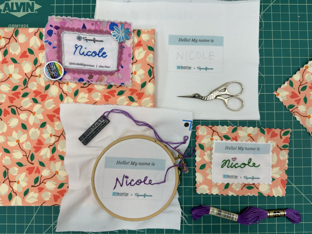 Several embroidered name tags with the name Nicole lay on a green cutting mat. A finished name tag on the pink fabric with magnolias, with the name Nicole stitched in green and a red heart as a dot over the letter “I” in Nicole. A name tag stitched with the name Nicole in purple embroidery floss. A set of stork scissors are at the top of the photo, lying on white canvas.