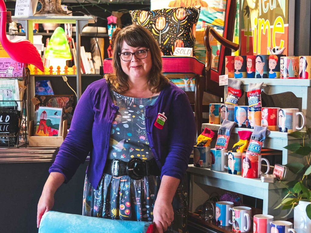 Kara stands in her studio behind a blue upholstered chair, wearing a purple cardigan over a dress made of her Australian Animals design on grey, featuring quolls, lyrebirds and kangaroos surrounded by illustrated Australian flowers and plants. Behind her are store shelves packed with colorfully illustrated pillows, socks, mugs, greeting cards and a plastic flamingo.