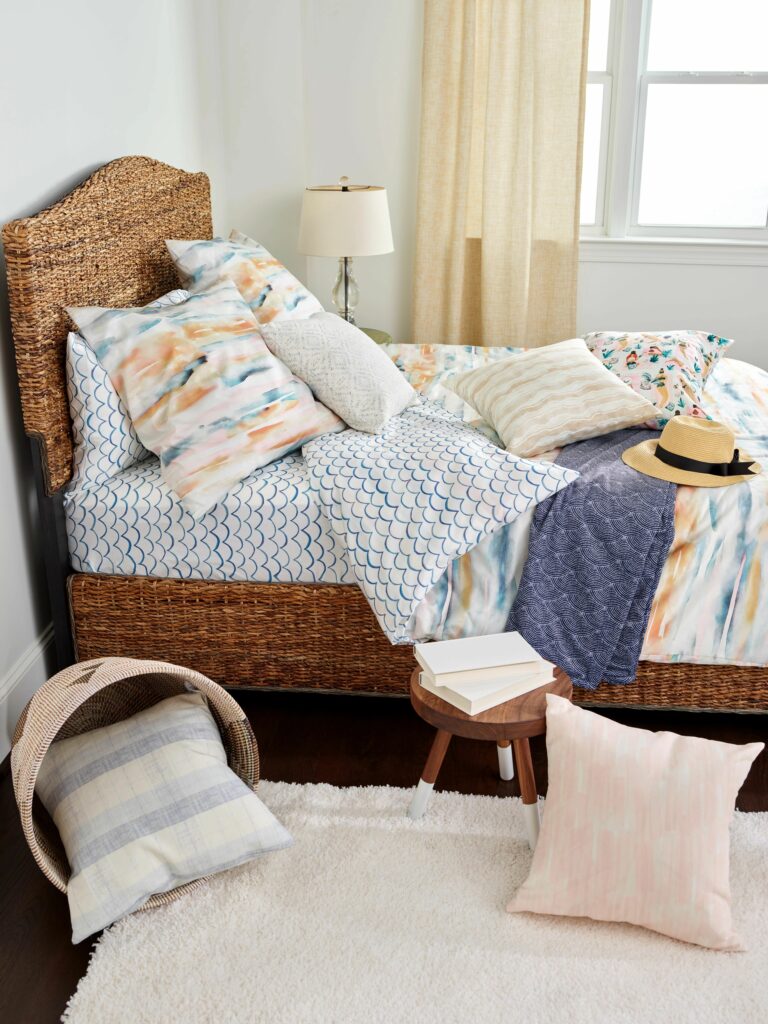 A bed with a rattan headboard and frame sits in front of a window with a cream curtain pulled to the left side. The bed is covered with linens that are white and blue and pastel.