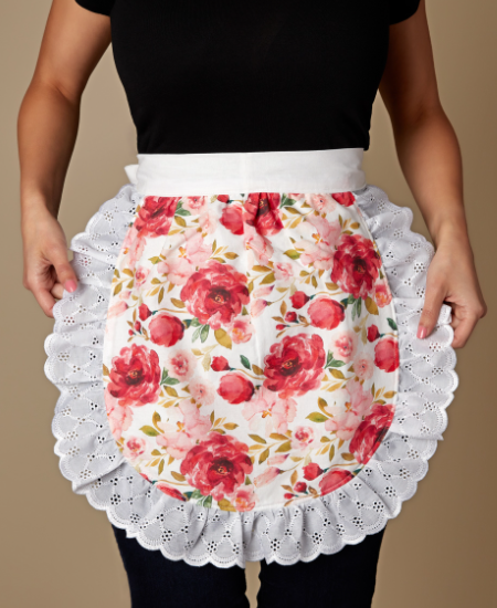 Image of a person wearing a floral apron; only their torso, arms and legs are in the photo. The apron is half-moon shaped and features a wide lace edging and a white fabric strip at the top and has a repeating red and pink floral design.