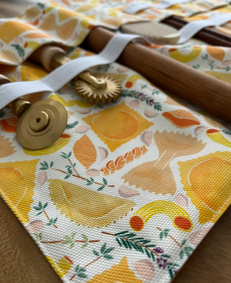 A close up of a fabric pasta tool case where tools for pasta making are held in place in small fabric pockets and secured at their tops with a strip of white elastic. The case’s fabric design features various repeating pasta shapes and small bits of herbs on a white background.