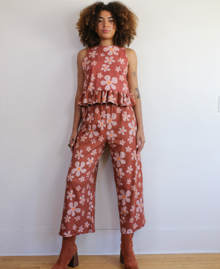 A model wears a matching floral two-piece set with cropped pants and a sleeveless peplum shirt. She stands looking at the camera against a white wall. The fabric design has repeating pink flowers in varying sizes with orange centers on a light burgundy background.