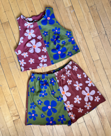 A matching tank top and shorts set has been laid out on a wooden floor. Both the tank top and shorts feature two similar but different floral designs. On the tank top, the left half of the fabric has repeating pink flowers in varying sizes with orange centers on a light burgundy background. The right half has blue flowers of varying sizes with pink centers on an olive green background. On the shorts, the left half of the fabric features the design with the blue flowers and the right half of the fabric features the design with the pink flowers.