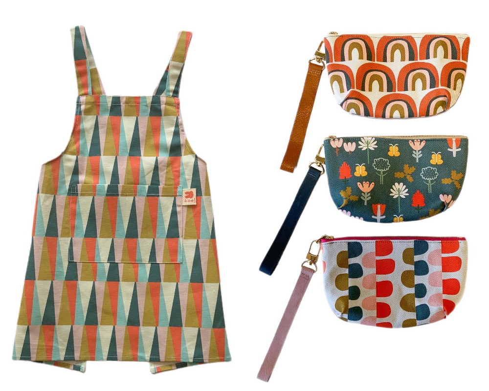Two images have been placed into one rectangular image. On the left, a children’s apron against a white background. The apron has a front pocket at the top and has rows of repeating long thin triangles in white, light blue, dark blue, pink and olive green. On the right, three small half-moon shaped clutch purses with straps hanging down and to the right against a white background. The top purse has repeating rows of muted pink, dark blue and orange rainbows. The middle purse has red, white and pink flowers as well as yellow butterflies and green leaves on a dark blue background. The bottom purse has vertical rows of repeating two half moons that meet in the middle of each row. The half moons are olive green, dark blue, red and pink. In each row, the half moon on the left side is a different color than the half moon on the right.