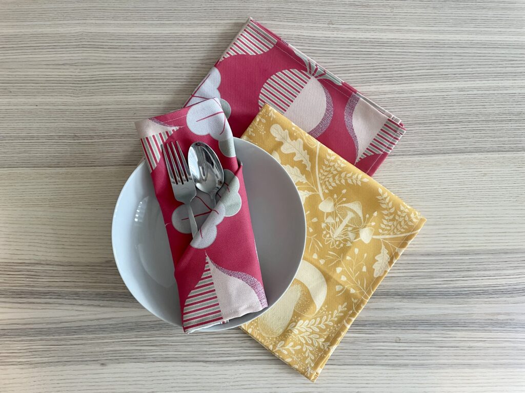 A folded napkin sit on top of a white ceramic bowl with a silver fork and spoon tucked inside. Two napkins lay tesselated underneath the bowl. The napkin folded on the bowl and the napkin laying directly against the table have a deep magenta background and repeating design with beets in a half drop repeat that are pink on one half, pink, magenta and gray striped on the other half and have gray tops. The napkin directly underneath the bowl has a deep yellow background and cream-colored mushrooms and small leaves. The bowl and napkins are lying on a light wooden table.