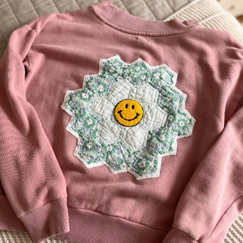 A dark dusty rose sweatshirt lies on a light pink sofa. A quilt block that is cream in the middle and green with a light floral print around the edges has been sewn to the front of the sweatshirt. The quilt block has a yellow smily face patch sewn in the middle of it.
