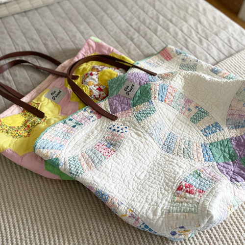 Two tote bags with dark leather handles lay pointing to the left on a cream sofa and blanket. The totes are made from reclaimed quilts. The tote bag on the bottom is yellow, pink and white. The tote bag on the top is mostly white but small floral sections from a double wedding ring quilt design run through the bag’s surface.