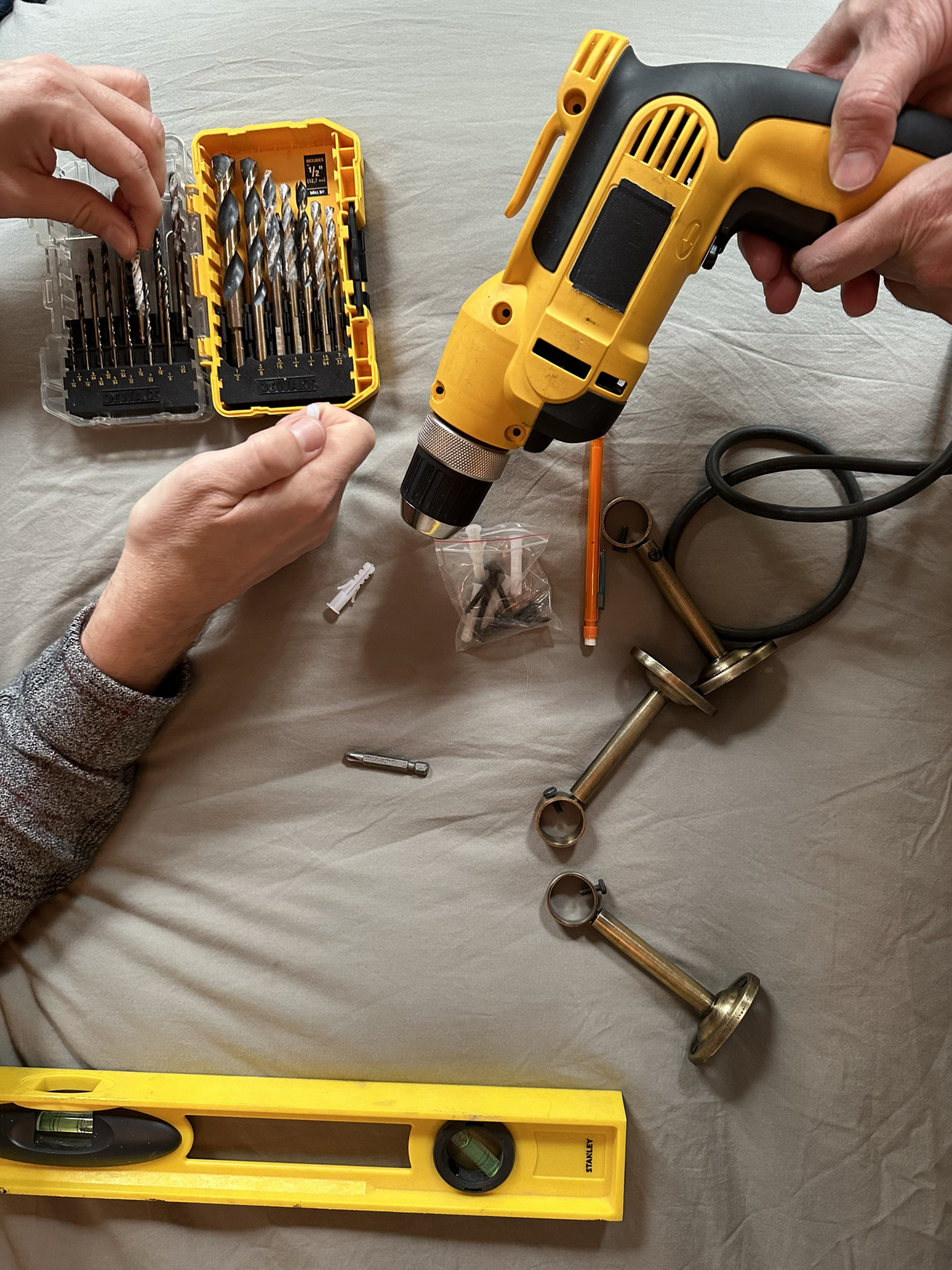 A yellow spirit level partially extends into the bottom left of the photo. Curtain installation hardware, drill bits, a pencil and a drill lay on a light gray sheet on a bed. In the top left, a hand is going through a container of drill bits. In the top right, another hand from another person holds a yellow drill.