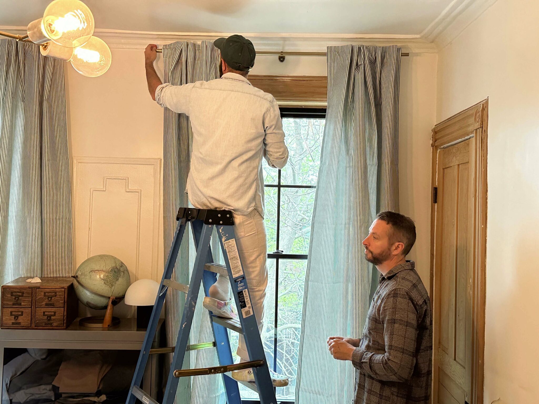 A man stands on a ladder and hangs up the left curtain panel of a pair of top tab dark blue-and-white-vertically striped curtain panels. The right panel has already been hung. Another man stands to the right assisting with the installation. A small bookcase sits to the left of the window; it has a small dome lamp, a painting of a man looking pensive and a globe on top of it.