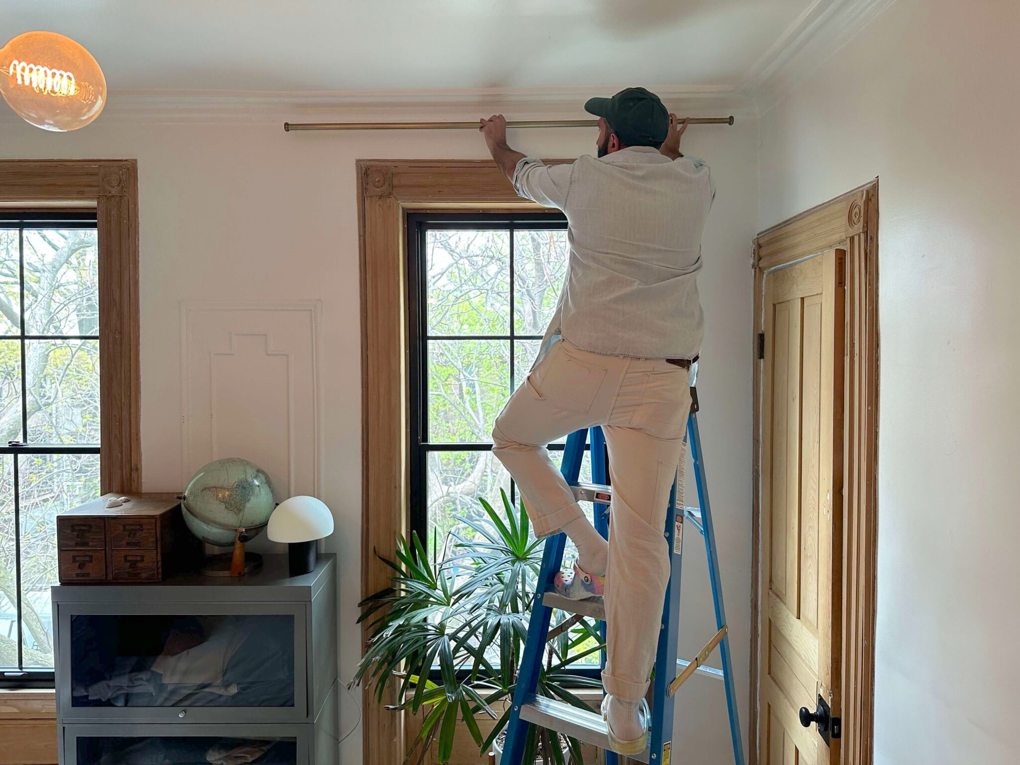 A man installs a metal curtain rod above a window with brown wooden molding. He is standing on a ladder.