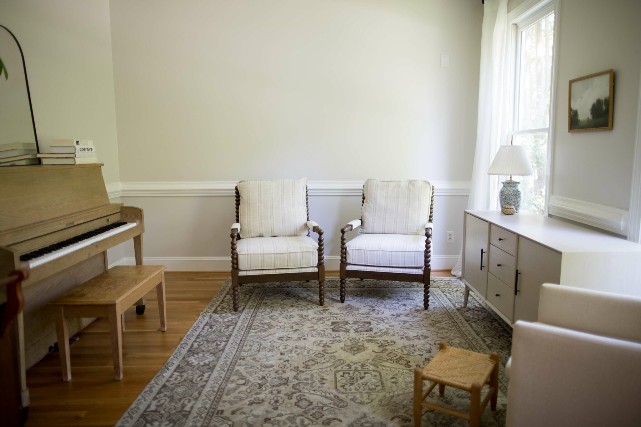 An image of the sitting room before the makeover. Two brown chairs covered in white cushions are to the image’s background. A white low storage table sits to the right and a piano sits to the left. The walls are cream.