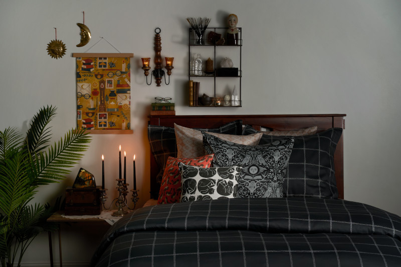a shadowy bedroom scene featuring black, plaid bedding, black pillar candles that are lit, and a green palm tree on the left side. There is a black wire shelf above the bed.