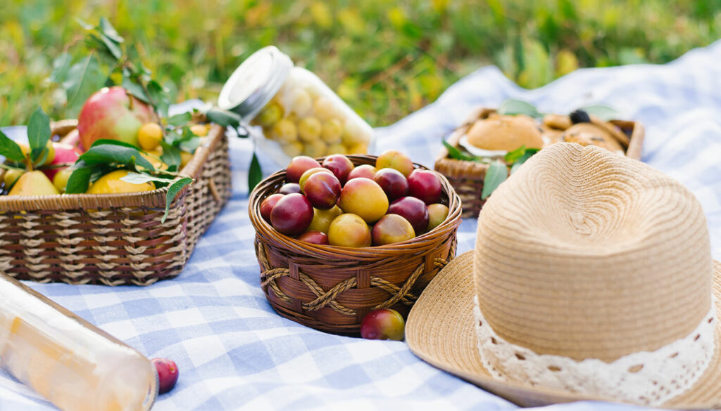 A close up image of a picnic set up on the grass. Baskets are filled with fruit and baked goods, a jar of olives and a straw hat rest on a light blue and white gingham tablecloth.