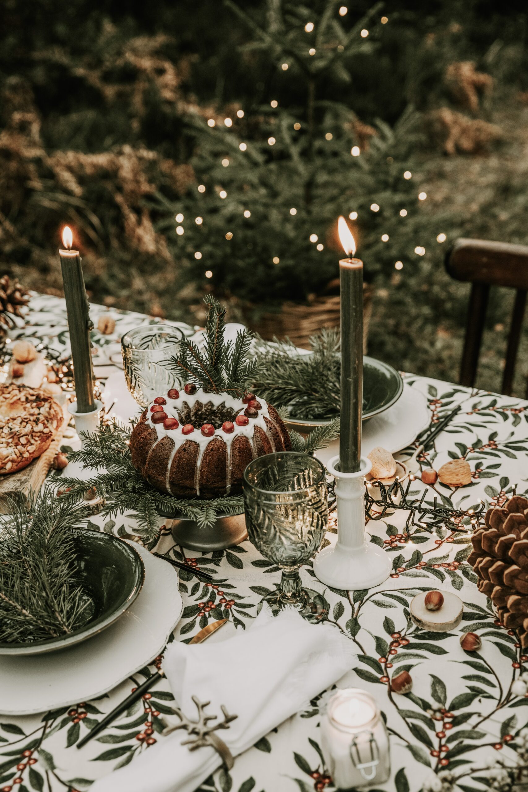 Image of a winter solstice tablescape with a cake with white icing and red fruit and lit dark green candles on a white tablecloth with a green leafy vine design. A green tree with small circle lights is in the background.