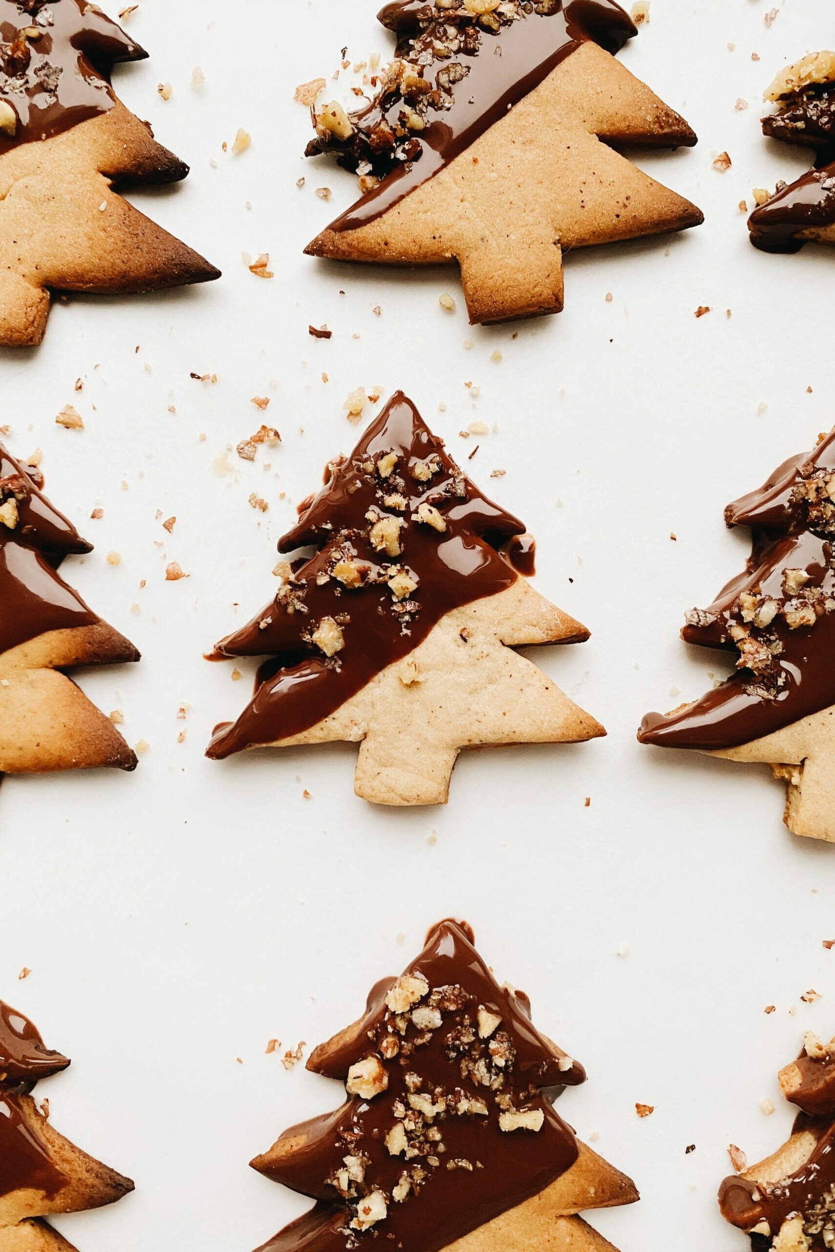 Rows of fir tree cookies lay on a white surface. They are partially covered in chocolate on the diagonal on the top left and sprinkled with small pieces of nuts.