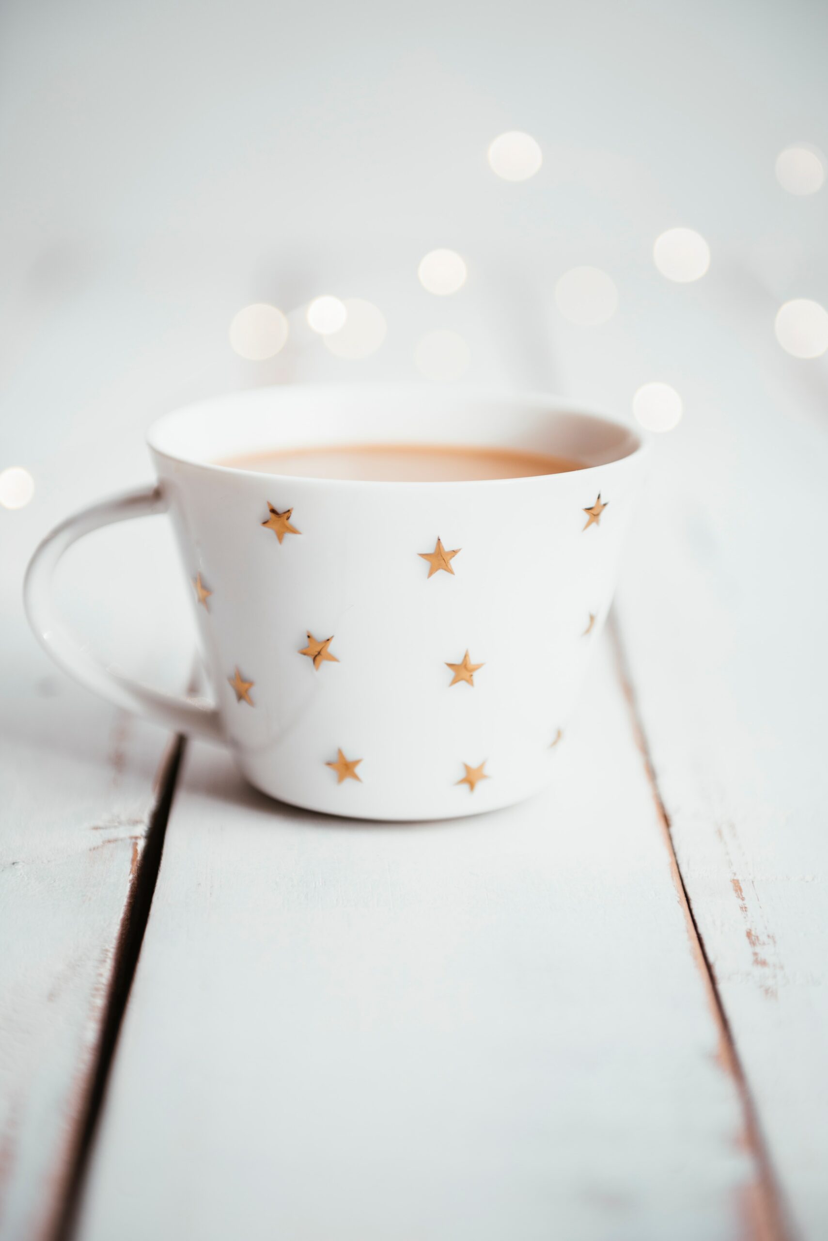 Image of a white mug with repeating gold stars on a white table. Small white lights are to the middle right.