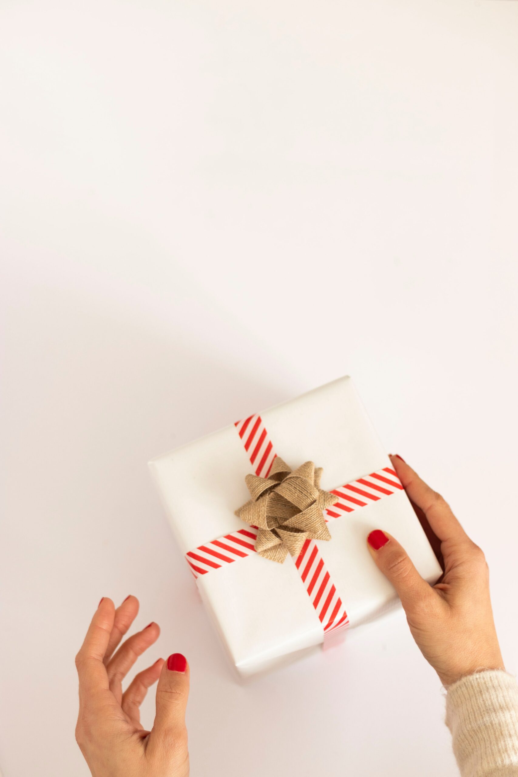Two hands reach out for a present laying on a white surface and as seen as shot from above. The present has white wrapping paper with a red-and-white trim wrapped around it and a light brown bow at the top.
