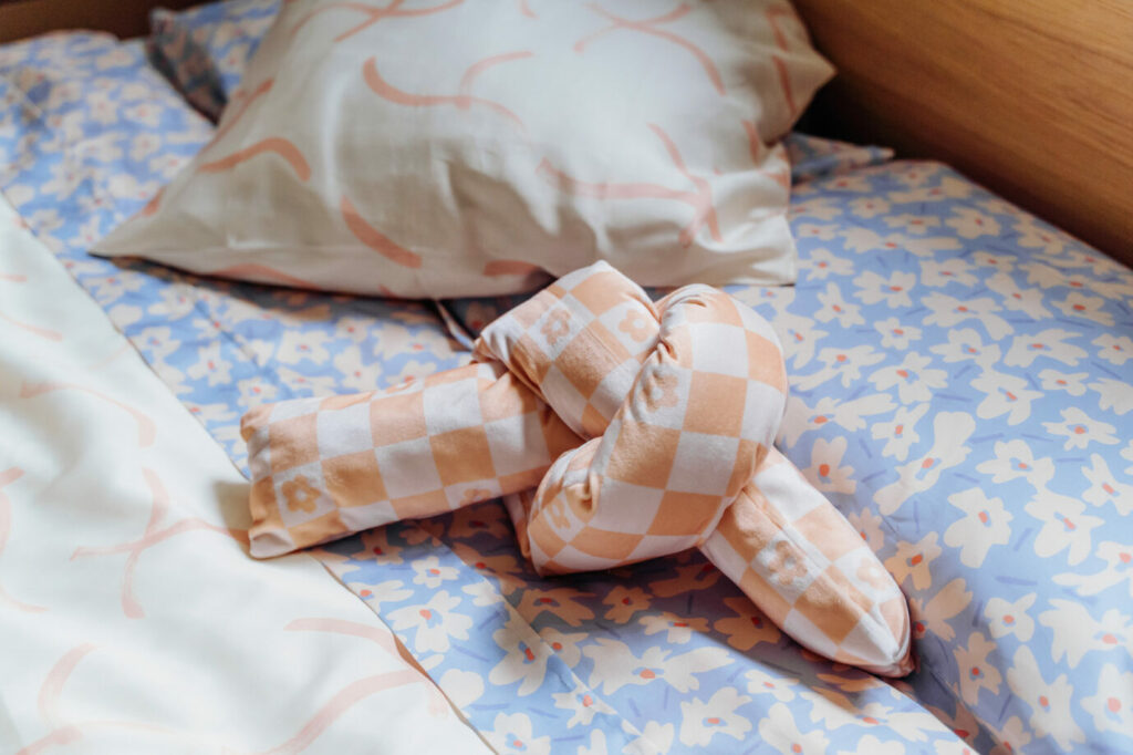 Orange and white knot pillow sitting on bed with purple flowered sheets and pillowed cases, and crème pillowed cases matching the crème duvet.