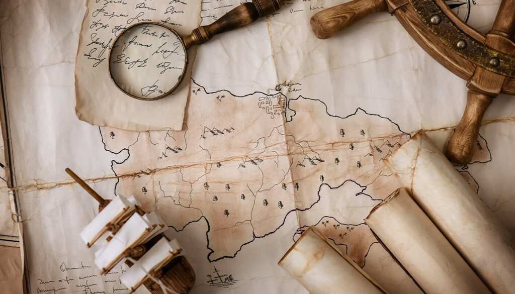 From a bird's eye view you see a vintage hand drawn map, stained and wrinkled, spread across a table. On top of the map sit a handwritten note with torn edges, a magnifying glass, a wooden ship’s wheel, three rolled up maps and a small model ship.