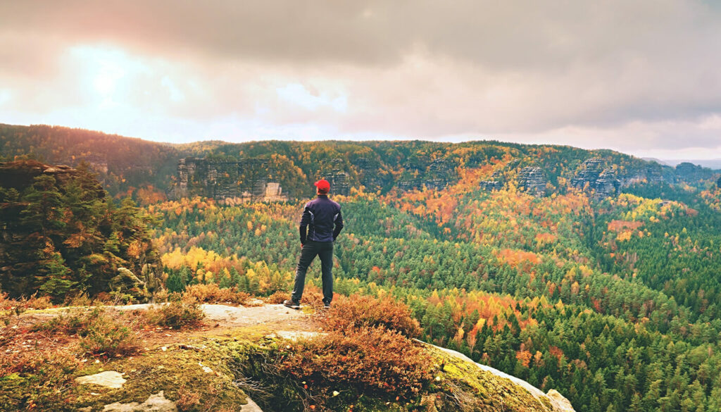 A man stands at the top of a mountain with his back turned to us, looking out at the view of mountains covered in trees with leaves beginning to change to beautiful hues of red, orange and yellow signaling the beginning of autumn.