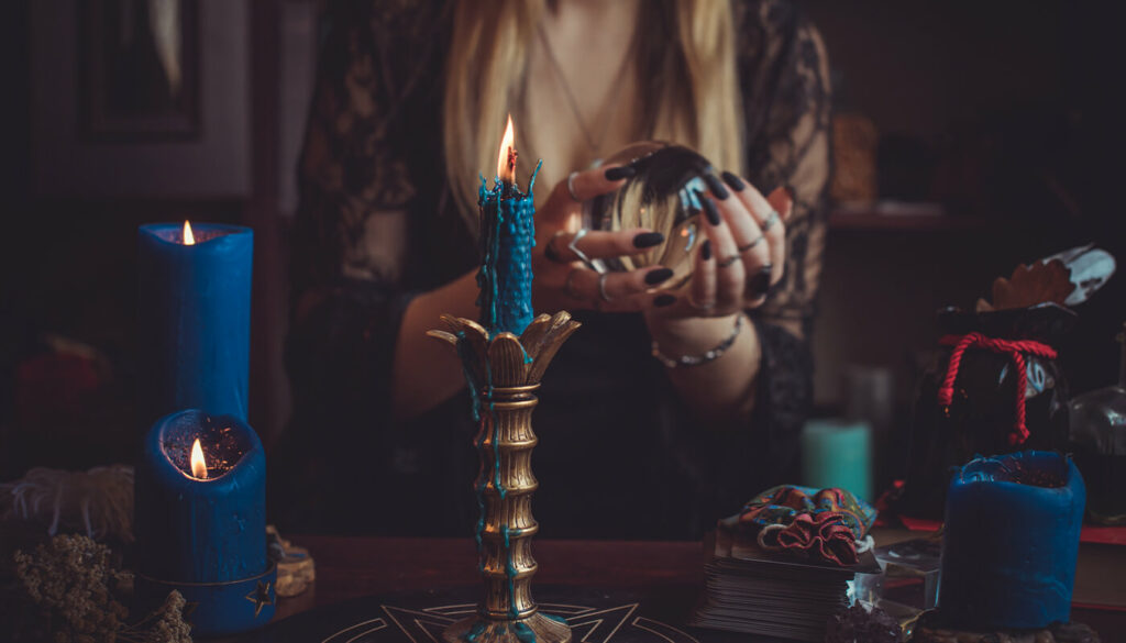 The image is cropped in on a dark room, focused on a woman’s hands with long black nails and stacked silver rings holding a clear crystal ball. In front of her are blue candles burning with wax dripping down the sides, a deck of cards, dried flowers and small bags tied with red cord. 