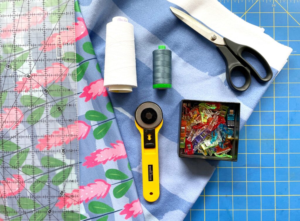 Materials to create a fleece headband are spread out across a blue and yellow cutting mat. A blue-striped fleece fabric is under a black pair of scissors, a white spool of thread and a forest green spool of thread and a rotary blade. A striped fabric with pink flowers and green leaves lays between the materials, and underneath a clear cutting mat.